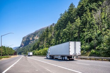 Convoy of big rigs semi trucks with semi trailers moving on the highway road with forest and mountain on the side