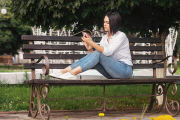 young beautiful brunette girl in a white shirt and blue jeans sits on the street in the park drinks coffee from a glass and looks through her phone