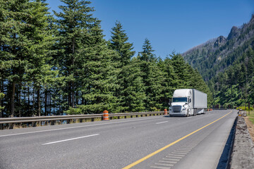 White industrial big rig semi truck with dry van semi trailer transporting cargo running on the scenic highway road with trees and mountain in Columbia Gorge area