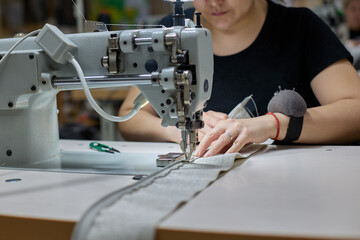 Seamstress or an employee of an Asian textile factory sewing on an industrial sewing machine. Close-up of the process of sewing fabric by a woman in production - obrazy, fototapety, plakaty