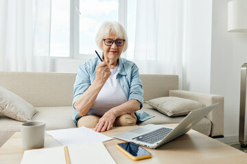 an enthusiastic, happy, emotional elderly woman works from home sitting on a bright sofa in her apartment with a laptop on a coffee table and emotionally raises her hand up