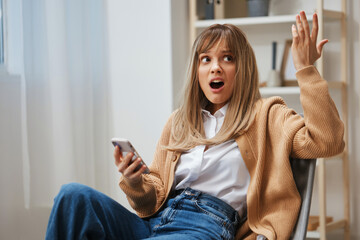 Shocked confused unhappy young blonde lady in warm sweater open mouth raise hand up hold phone sitting in armchair at home. Pause from work, take a break, social media in free time concept. Copy space