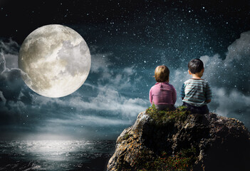Little boy and girl sitting on a rock above the sea looking at the moon and stars