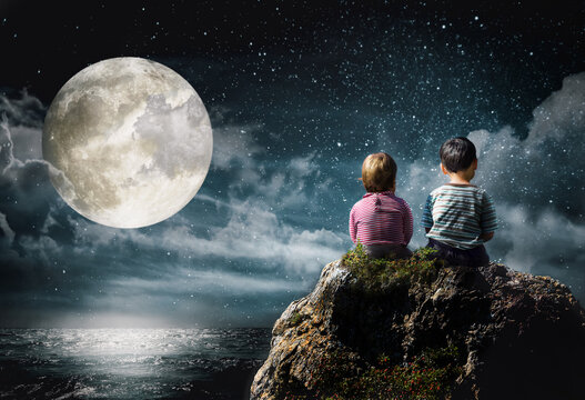 Little boy and girl sitting on a rock above the sea looking at the moon and stars