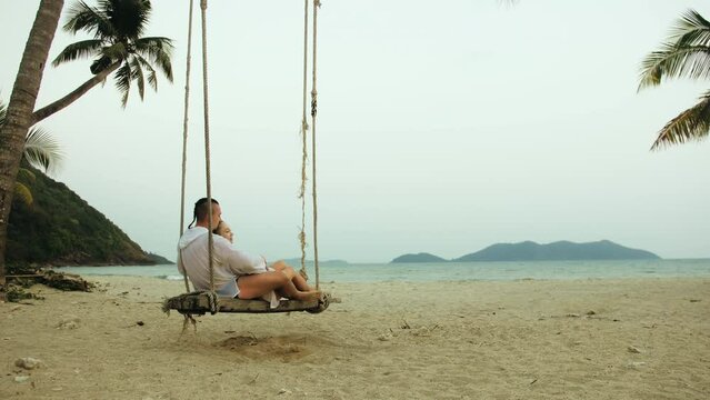 Cute loving couple on the swing on tropical beach. Woman and man in white shirt enjoy gold sunset and life in beaches, palm tree. Evening warm sunset. Summer holiday vacation tropical tourism concept
