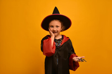 Cute little wizard girl holding toy spider and looking at camera on a yellow background.

Surprised...