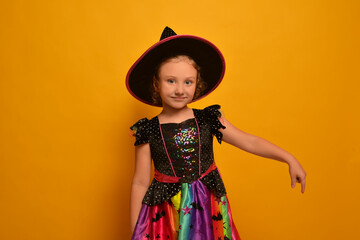 Little cute girl in witch costume looking at camera and pointing index finger down or holding finger down on a yellow background.

Joyful wizard child wearing Halloween dress and hat.