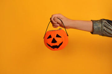 Orange pumpkin candy bucket or Halloween Jack o Lantern candy holder with sweets in female child hand on a yellow background with copy space.

Happy Halloween concept. - obrazy, fototapety, plakaty