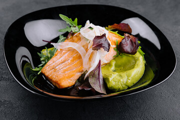 Close-up of grilled salmon fillet with green arugula sprouts with mashed avocado on a plate.