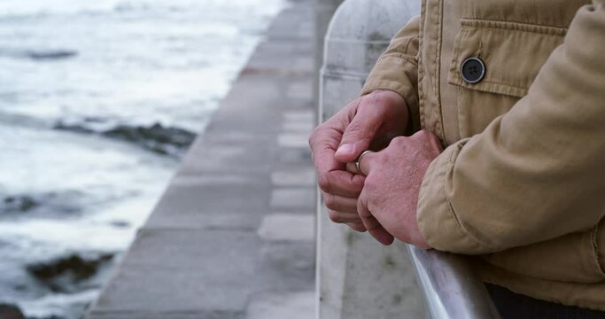 Widower feeling alone and sad on a walk. Closeup of a mature man holding his wedding ring while standing at a beach on a cloudy day. Grieving man suffering from depression after losing his wife.