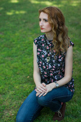 Red hair girl sitting on the grass. Beautiful girl wearing a floral shirt and jeans. Woman with curly long hair and makeup.