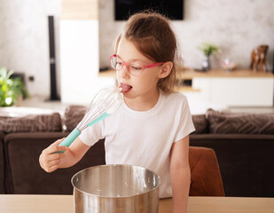 A cute Caucasian little girl licks a whisk with the remains of whipped raw cream, near the table in...