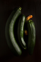 Three dark green zucchini with flowers in the dim light on the wooden table, flat lay