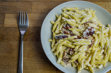 strozzapreti cream and speck, specialty from northern Italy, fresh twisted pasta with sauce made of fresh cream and speck, a smoked ham, and parmesan - in a white plate on a wooden cutting board