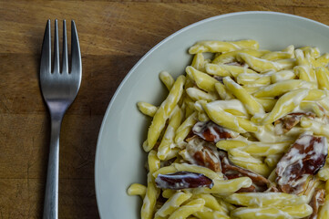 strozzapreti cream and speck, specialty from northern Italy, fresh twisted pasta with sauce made of fresh cream and speck, a smoked ham, and parmesan - in a white plate on a wooden cutting board