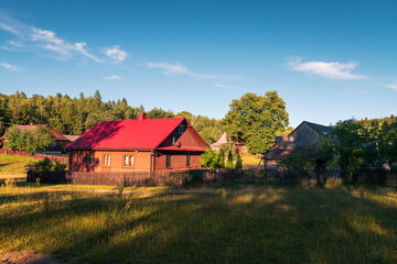 Fototapeta na wymiar Beautiful countryside settlement in the light of the afternoon sun. A house with a red roof and a wooden fence in the foreground, Krasnobród, Roztocze, Poland