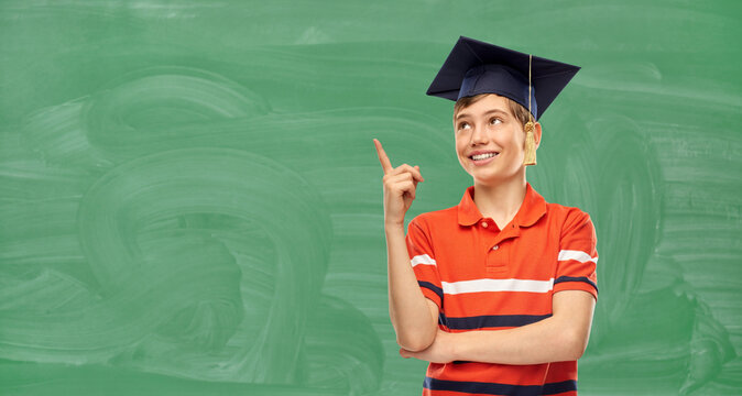 school, education and graduation concept - portrait of happy smiling graduate student boy in bachelor hat or mortarboard pointing finger up over green chalkboard background