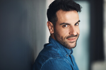 Portrait of confident and casual man leaning against a grey wall looking happy, proud and glad while smiling. Face of stylish, handsome and young european male standing alone inside with copy space