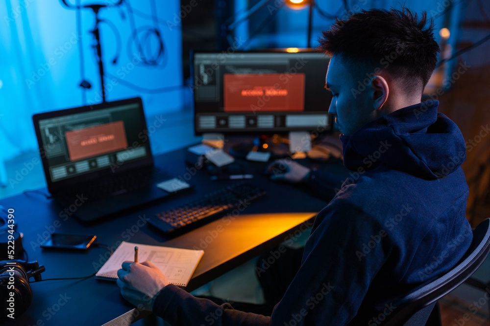 Poster cybercrime, hacking and technology concept - male hacker in dark room writing to notebook and using 