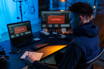 cybercrime, hacking and technology concept - male hacker in dark room writing to notebook and using...