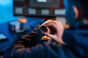 cybercrime, hacking and technology concept - close up of male hacker in dark room with smart watch and computer carries out cyber attack in dark room