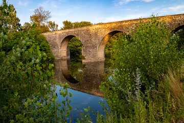 Fototapeta na wymiar Old roman bridge spanning over Cèze river in Provence south France near idyllic village La Roque and popular “Cascades du Sautadet“. Evening atmosphere with brick arches reflected on water surface.