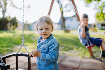 Little sibling playing together in playground, swaying on a swing, enjoying summer day.