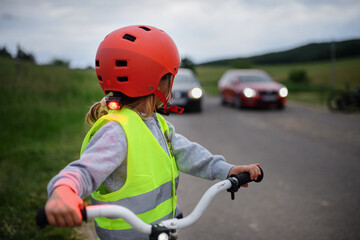 Rear view of little girl in reflective vest riding bike on road with cars behind her, road safety...