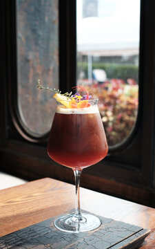 Black Coffee Shake In A Tall Glass On A Table In A Coffee Shop Or Cafe. Close Up Look Of Americano Shake In A Wine Glass Decorating With Lemon, Herbs And Flowers. A Modern Cafe Drinks With Copy Space.