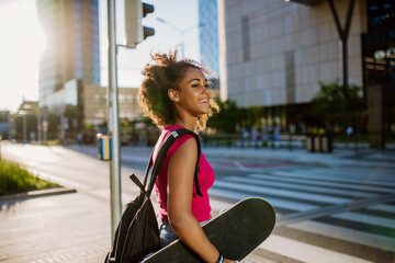 Multiracial teenage girl with backpack and skateboard, walking in city during summer day. - Powered by Adobe