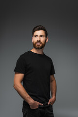 portrait of pleased bearded man in black t-shirt posing with hands in pockets isolated on grey.
