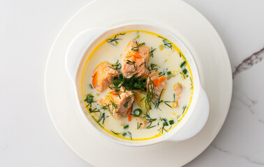 Norwegian red salmon fish soup with vegetables, herbs and cream, top view of a bowl of soup on a white marble background