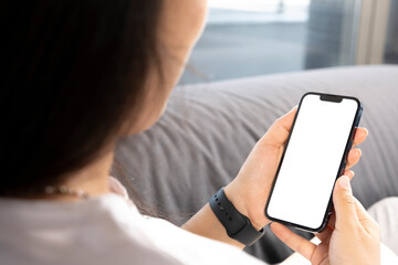 Holding smart phone, young woman sitting on sofa and holding smart phone with mockup white blank display. Vertical empty screen for social media apps. Over shoulder closeup view.