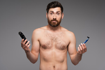 Fototapeta premium confused and shirtless man with beard holding safety and electric razors isolated on grey.