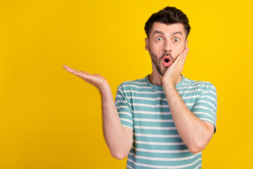 Photo of ecstatic impressed man dressed striped t-shirt demonstrating empty space hand on cheek isolated on yellow color background