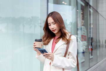 Obraz na płótnie Canvas Beautiful asian female businesswoman use smartphone hold paper cup of hot drink, Walk enjoy smiling while doing commuting in the modern city near office building outside