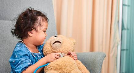 Cute curly haired Asian girl with stethoscope plays with teddy bear.
