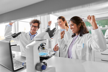 science, work and people concept - international group of happy smiling scientists with laptop celebrating success in laboratory