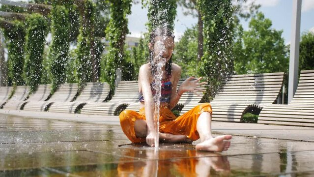 A preteen girl sits on the slabs of the city fountain in the park and plays with the fountain jets spouting from the ground.