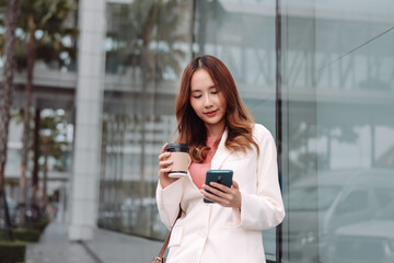 Fototapeta premium Beautiful asian female businesswoman use smartphone hold paper cup of hot drink, Walk enjoy smiling while doing commuting in the modern city near office building outside
