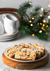 Fototapeta na wymiar Stollen fruit bread slices with dried fruit and marzipan coated with powdered icing sugar in the white plate on the table with Christmas tree and cups