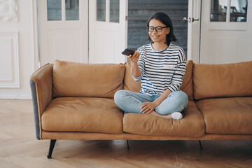 Smiling woman watching television series or movie changes TV channel, sitting on modern sofa at home