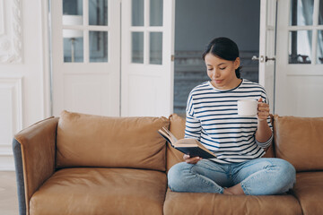 Woman rests reading bestseller novel holding paper book, coffee cup, sitting on sofa at home