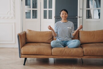 Obraz premium Smiling female practicing yoga, meditates, sitting on sofa at home. Healthy lifestyle, wellness
