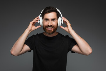 Naklejka premium cheerful and bearded man in wireless headphones listening music isolated on grey.