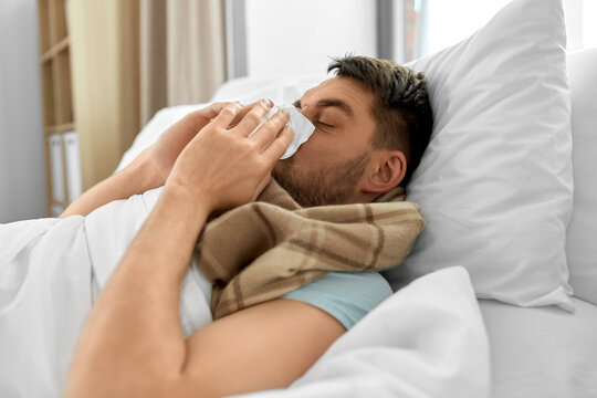 people and health problem concept - unhappy sick man blowing nose lying in bed at home