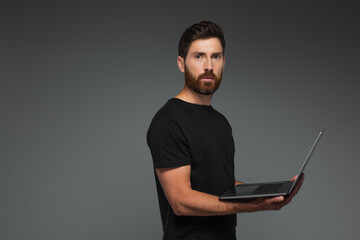 bearded freelancer in black t-shirt holding laptop isolated on grey.