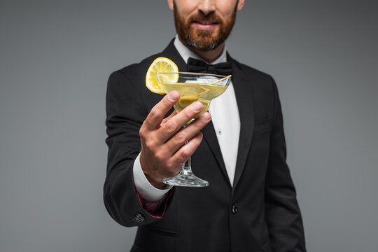 cropped view of cheerful and bearded man in suit with bow tie holding glass of cocktail isolated on grey. - Powered by Adobe