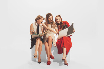 Naklejka premium Portrait of three young, beautiful, stylish woman sitting together and reading magazine isolated over white studio background