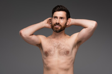 shirtless and happy man with hair on chest posing with hands behind head isolated on grey.